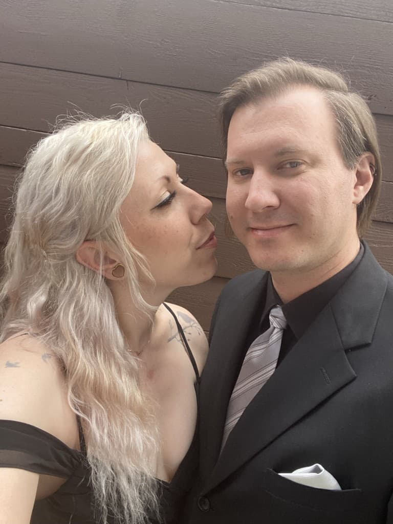 Dan and Madison in formal attire, Madison leaning in to kiss Dan on the cheek against a wooden wall