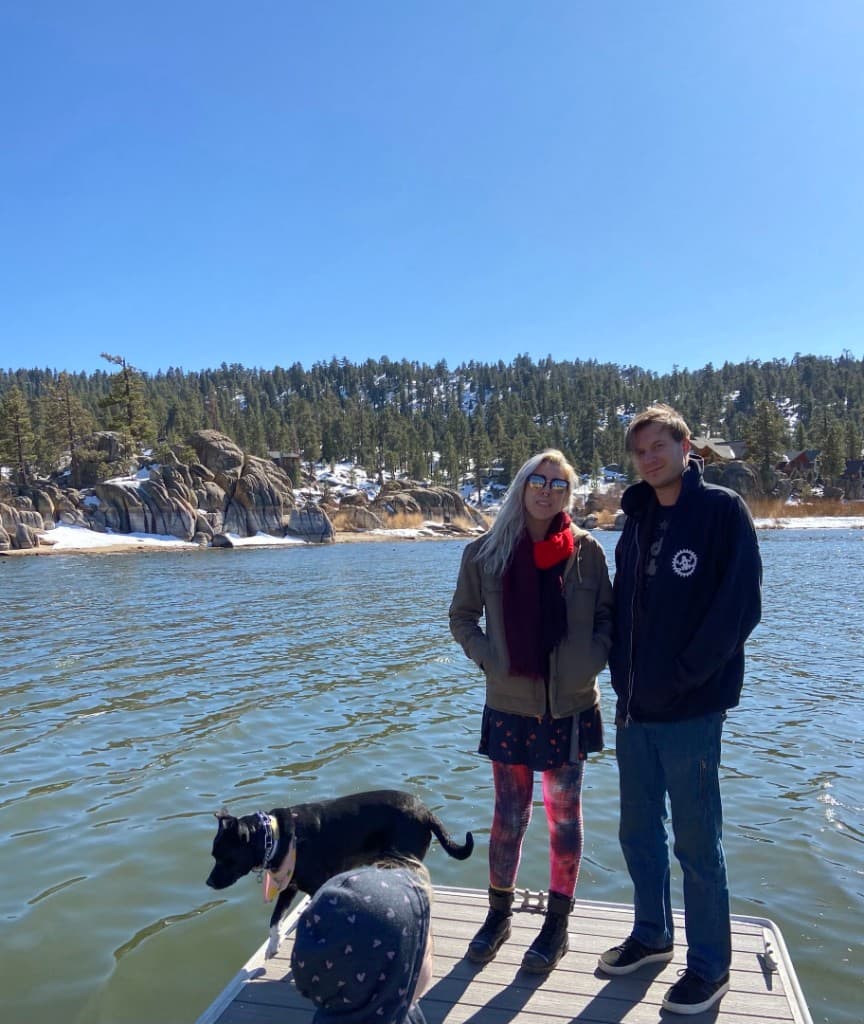 Dan and Madison with their dog Watson on a dock at the lake, mountains and pine forest in the background