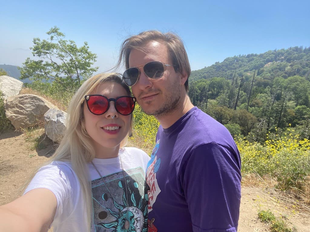 Dan and Madison on a sunny mountain trail selfie, green forested hills and blue sky behind them